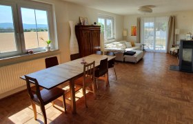 Helles Zimmer mit Esstisch, St&uuml;hlen, Sofa und gro&szlig;en Fenstern mit Blick auf die Landschaft.