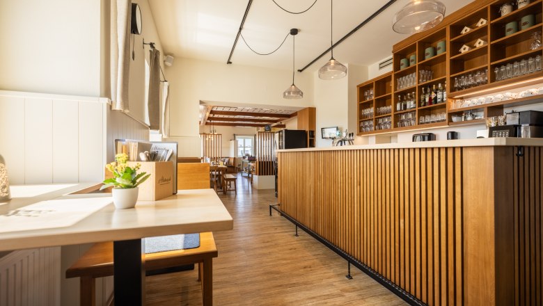 Innenansicht eines modernen Restaurants mit Holzmöbeln und Theke, durch Fenster kommt viel Licht herein, über der Schank hängen moderne Lampen.