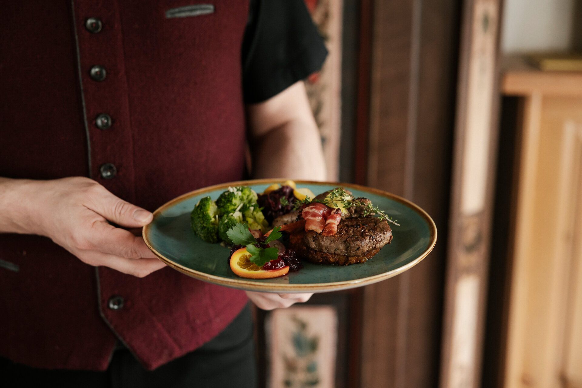 Angerichteter Teller mit Brokoli, Rindfleisch und Garnierung, gehalten von einem Mann in weinroter Trachtenweste.