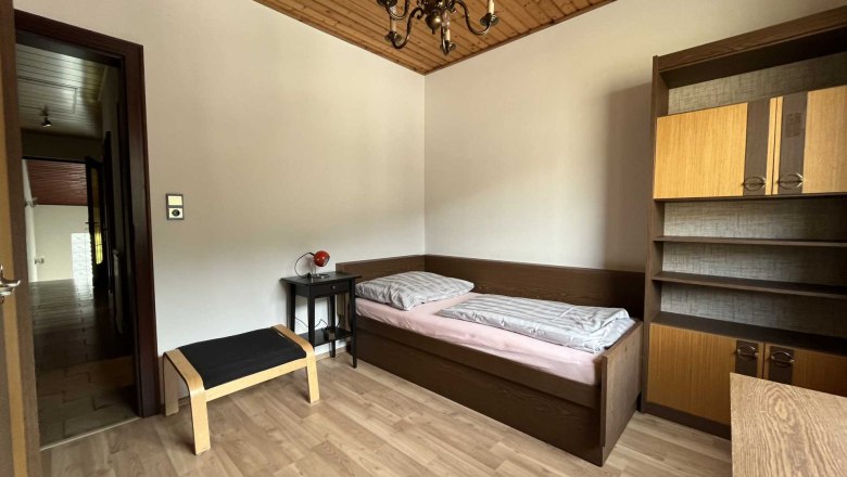 A simply furnished bedroom with single bed, wooden ceiling, shelf and small table with lamp.