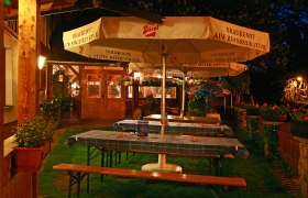 Ein gemütlicher Biergarten bei Nacht mit Holztischen, blau karierten Tischdecken und großen Sonnenschirmen der Marke Stiegl.