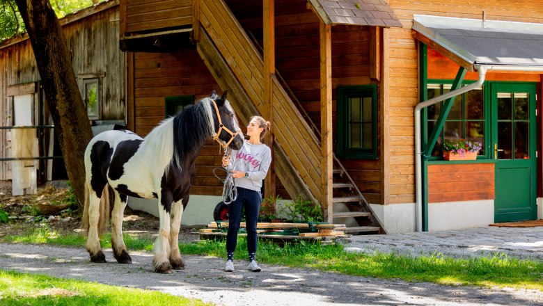 Eine Frau steht neben einem schwarz-weißen Pferd vor einem Holzgebäude.