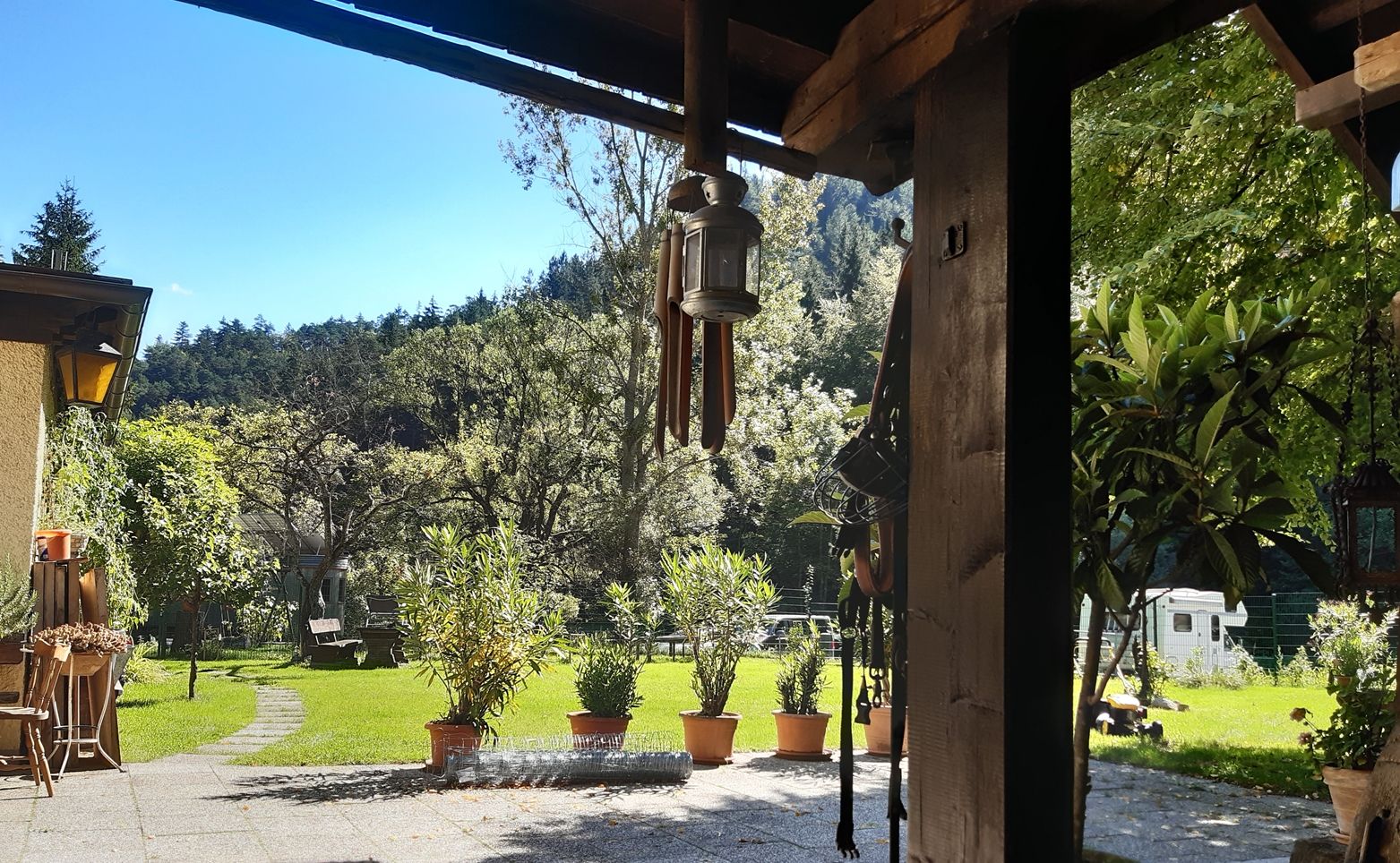 Ein sonniger Innenhof mit Rasen, Bäumen und Topfpflanzen. Im Vordergrund eine überdachte Terrasse mit einer Laterne und Gartenmöbeln.