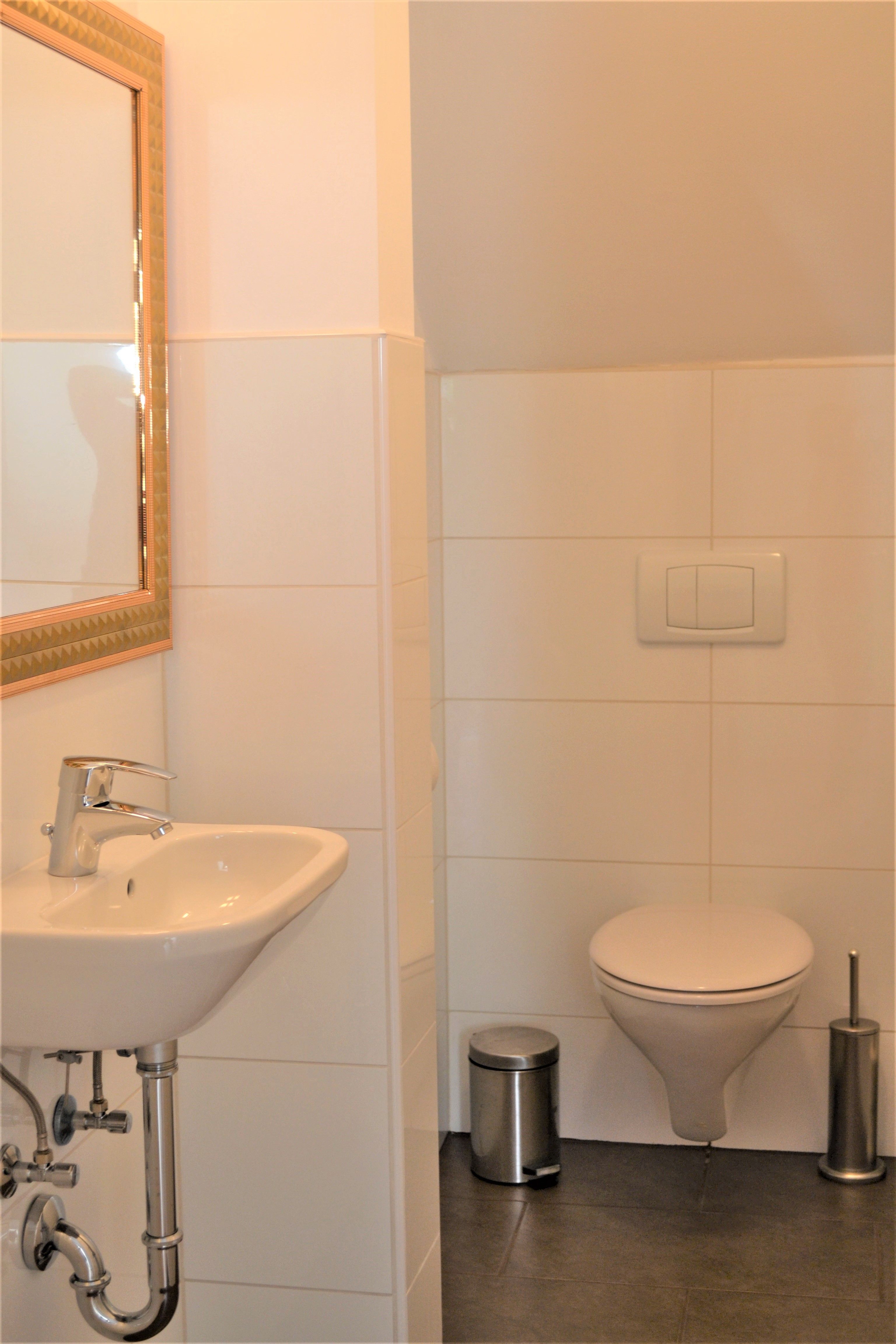 Kleines Badezimmer mit Waschbecken, Spiegel, Toilette, Mülleimer und Toilettenbürste.