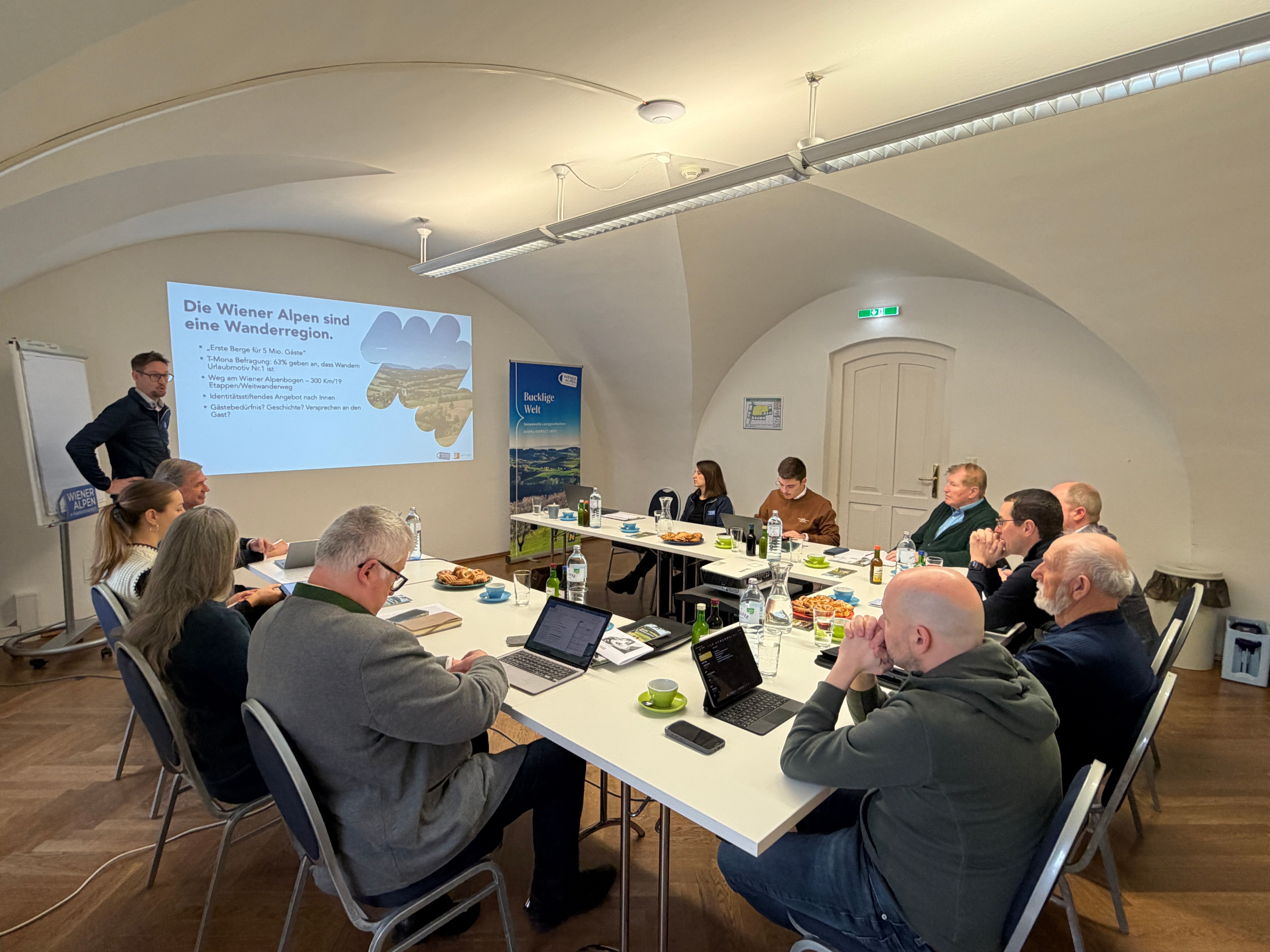 Mehrere Workshop Teilnehmer sitzen an einem Tisch während ein Mann einen Vortrag hält.