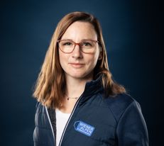 Portraitfoto Wiener Alpen Mitarbeiterin, die eine Jacke mit Wiener Alpen Logo trägt und vor einem dunklem Hintergrund steht.