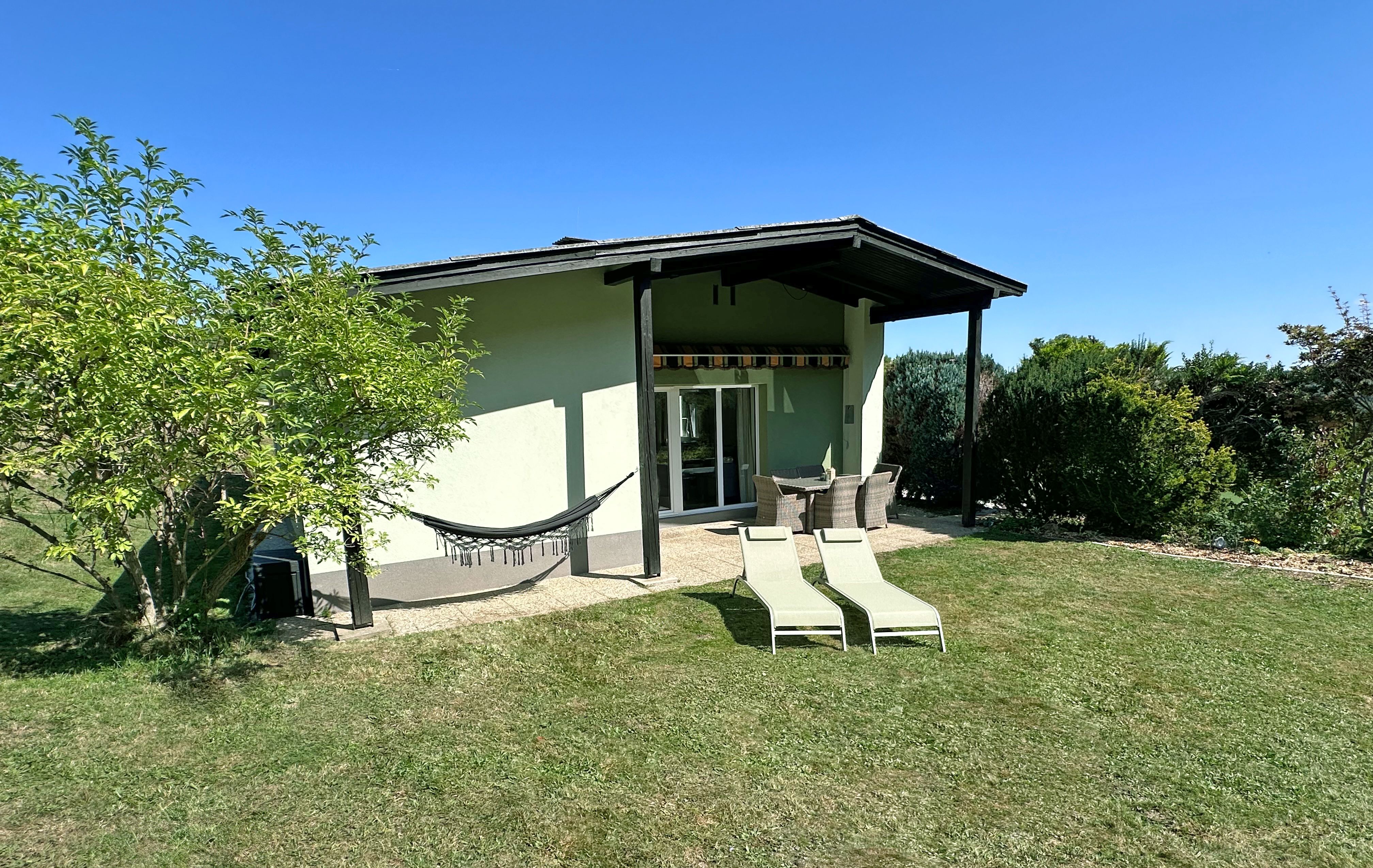 A small house with a patio, a hammock, two lounge chairs, and a garden under a blue sky.