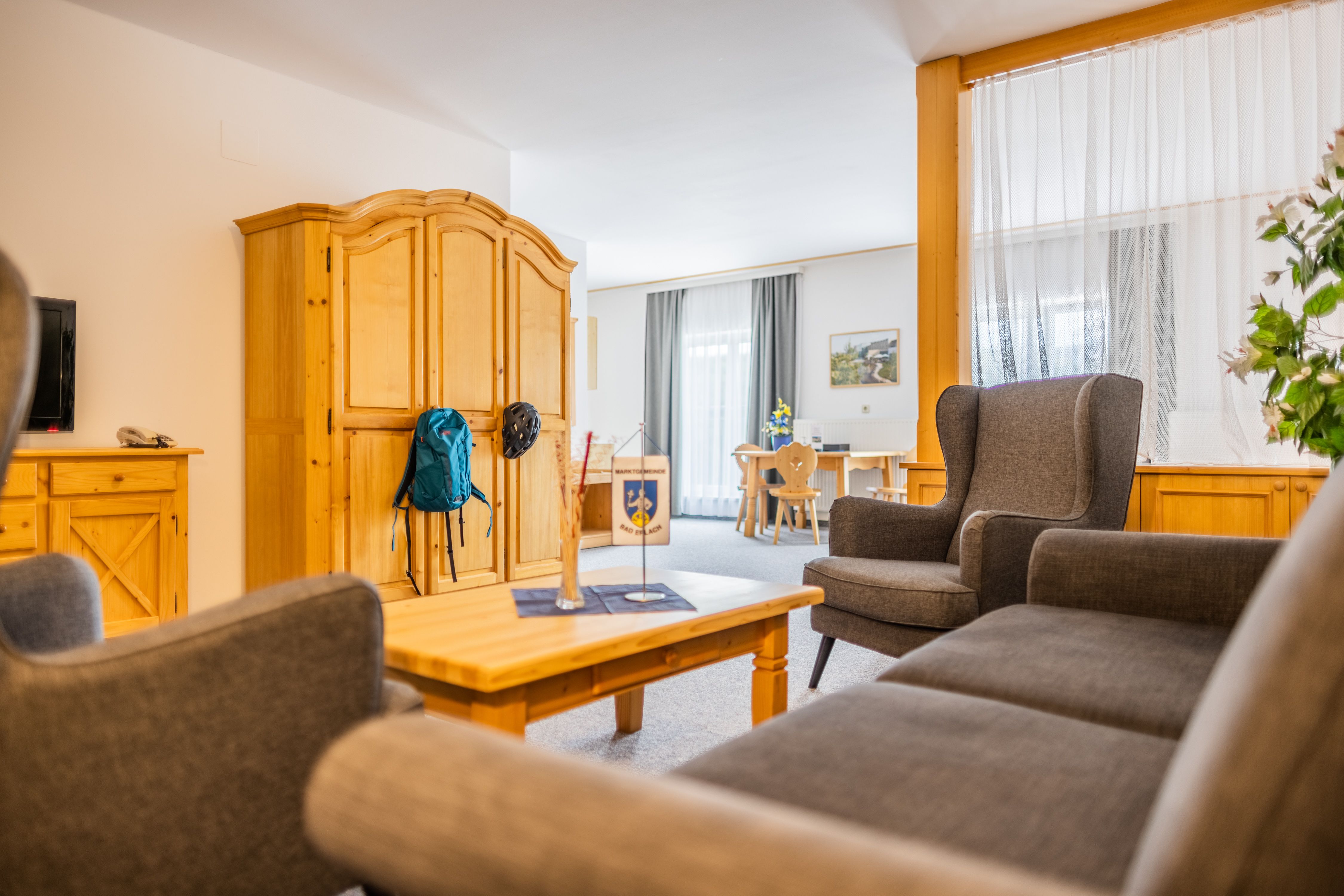 Gemütliches Wohnzimmer mit Holzmöbeln und grauen Sesseln, ein Rucksack hängt an einem Schrank.