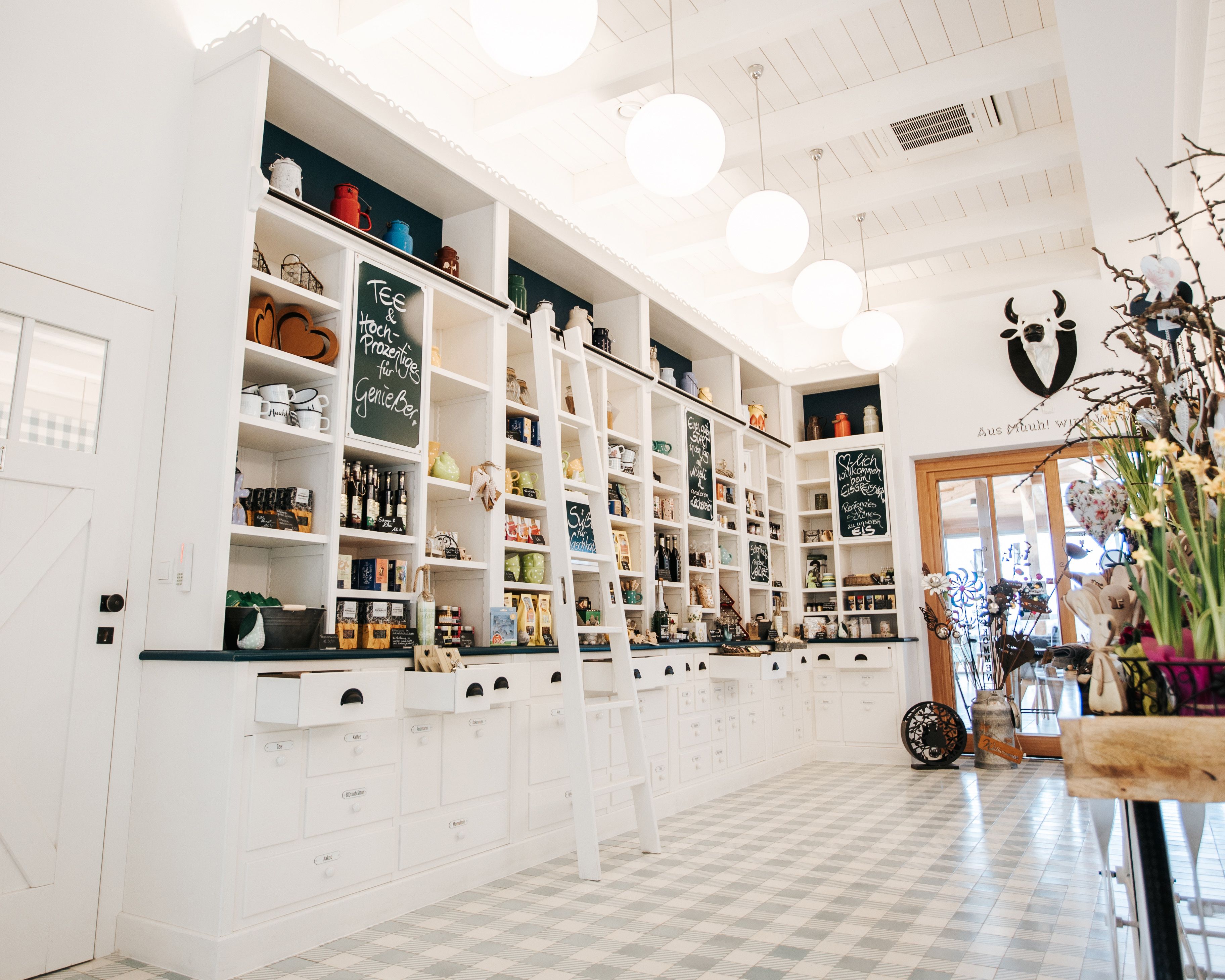 Innenansicht eines Hofladens mit weißen Regalen, kariertem Fußboden, regionalen Produkten und Dekoration.