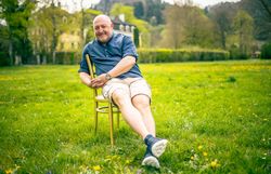 Mann sitzt entspannt auf einem Stuhl in einer blühenden Wiese und lächelt in die Kamera.