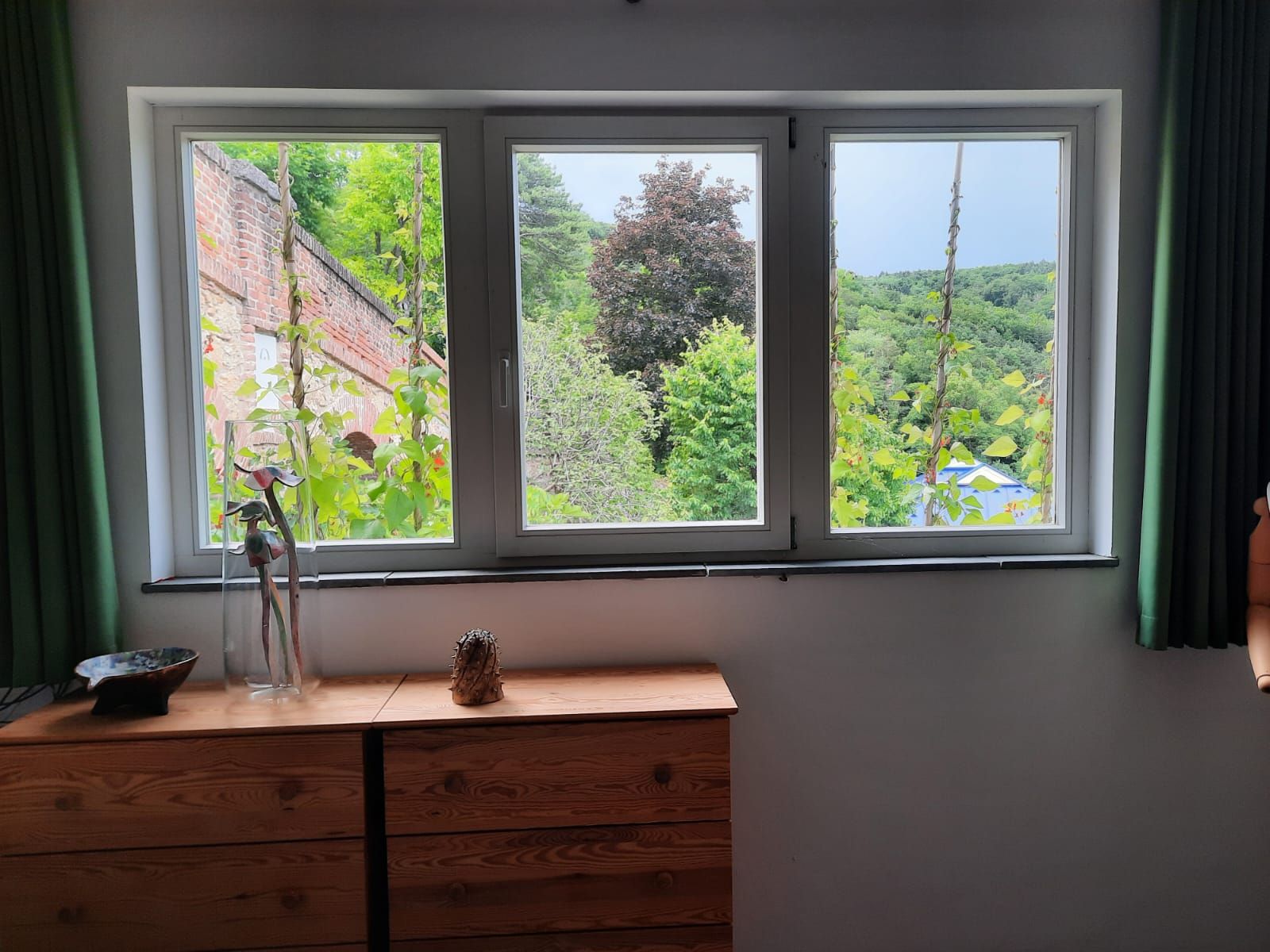 Blick aus einem Fenster mit grünen Vorhängen auf eine Landschaft mit Bäumen und einer Mauer.