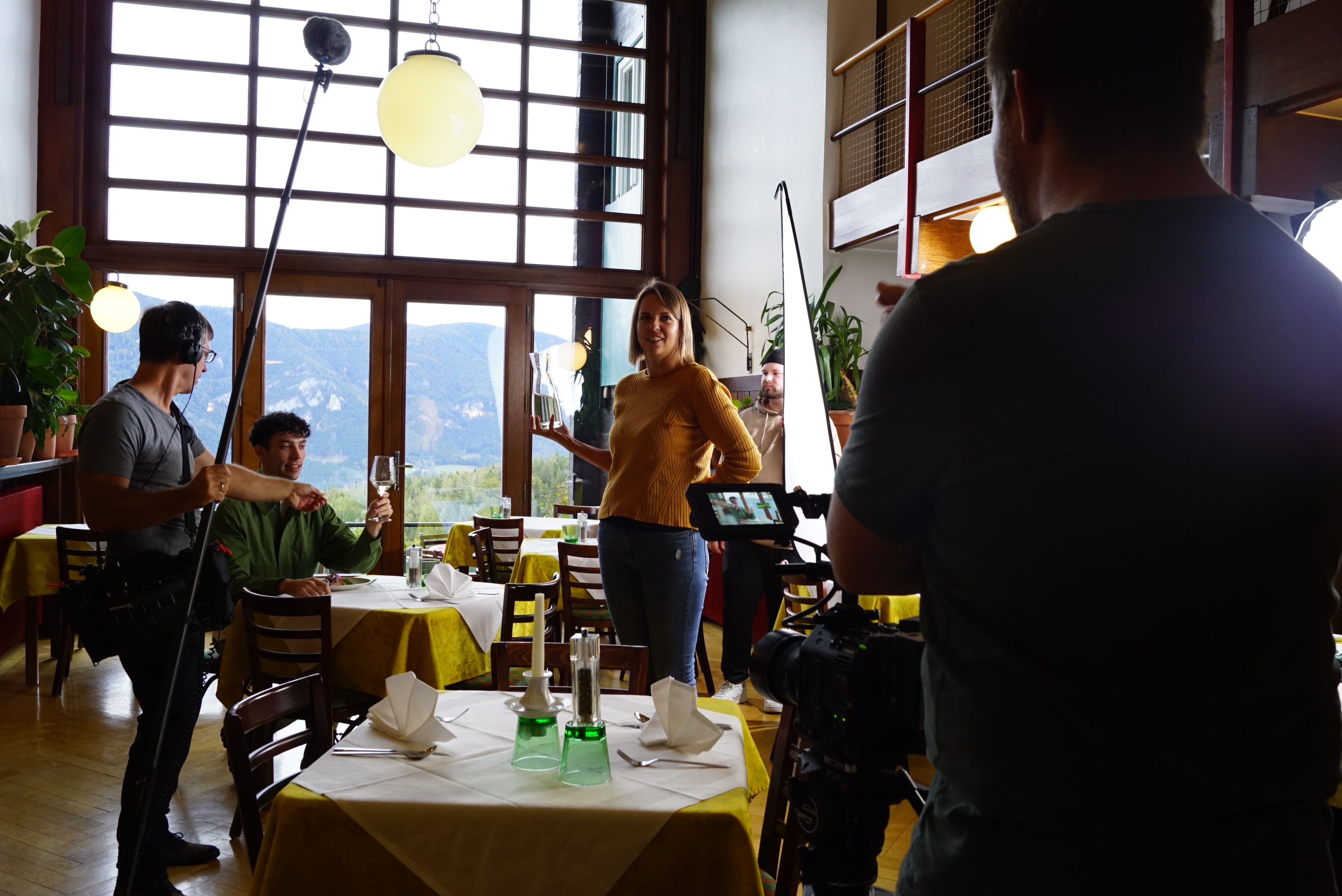 Bei Filmaufnahmen sitzt ein Darsteller in einem Gastzimmer mit Panoramafenster und Blick auf die Berge, an einem Tisch mit Glas in der Hand. Rund um ihn stehen Kameramann, Tonmann und eine weitere Frau mit Wasserkaraffe zum Nachschenken.