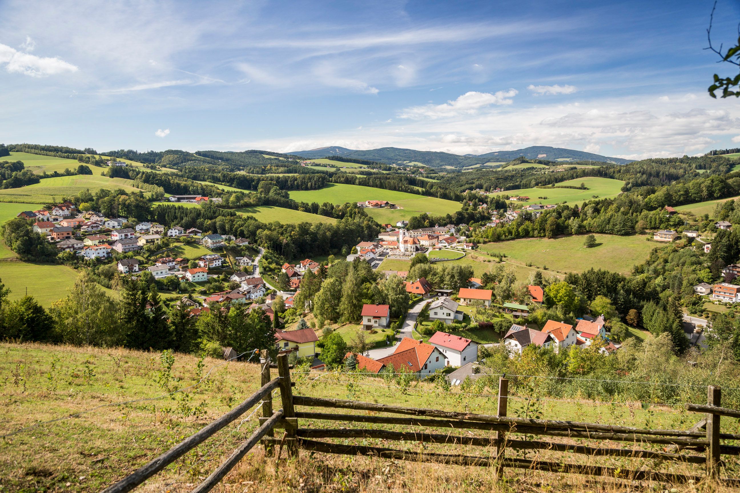 A view from a hilltop of Zöbern, a village with a church in the valley, surrounded by hills covered with meadows and forests.