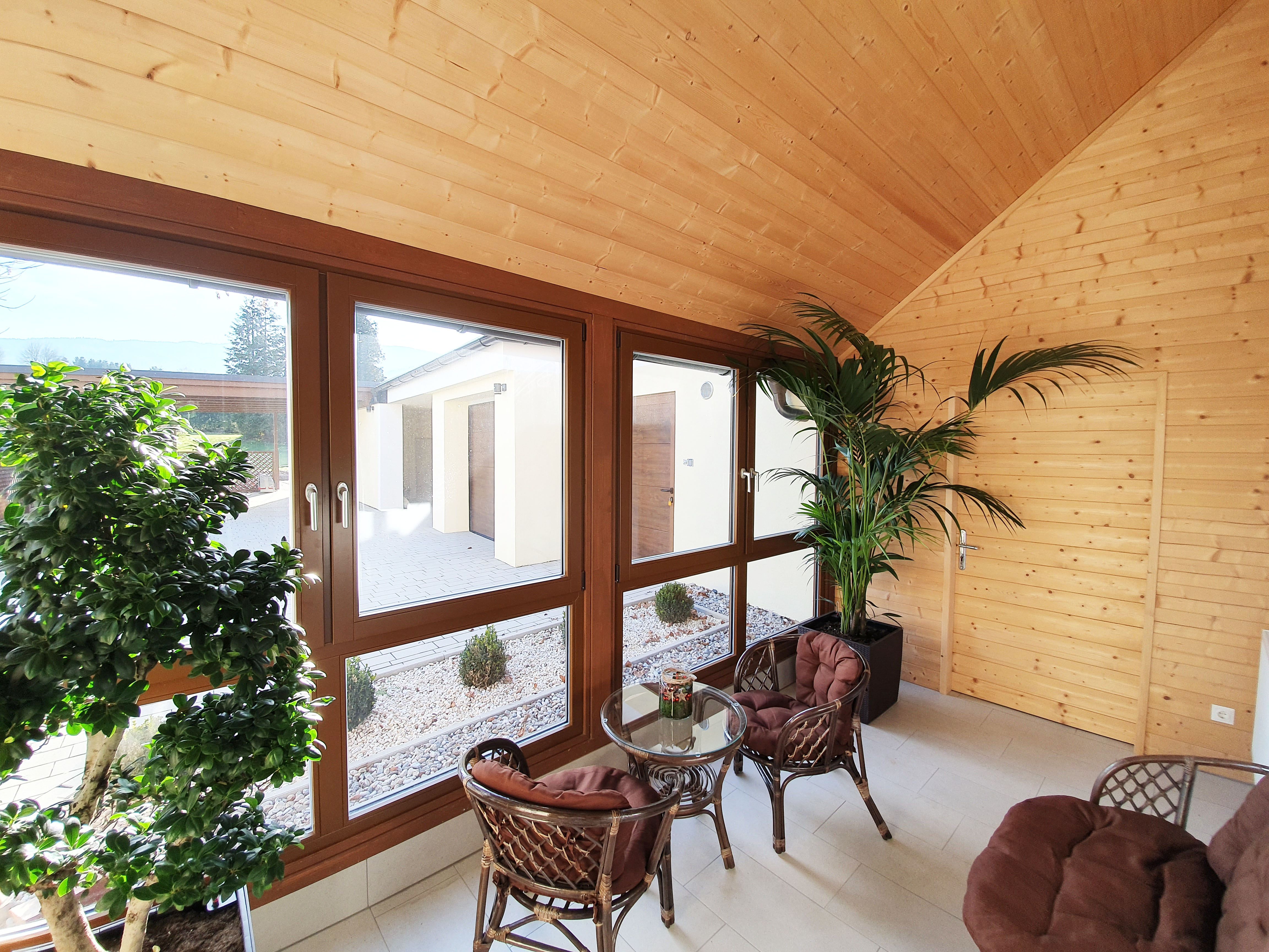 Innenansicht einer Veranda mit Holzwänden, großen Fenstern, Pflanzen und Rattanmöbeln.