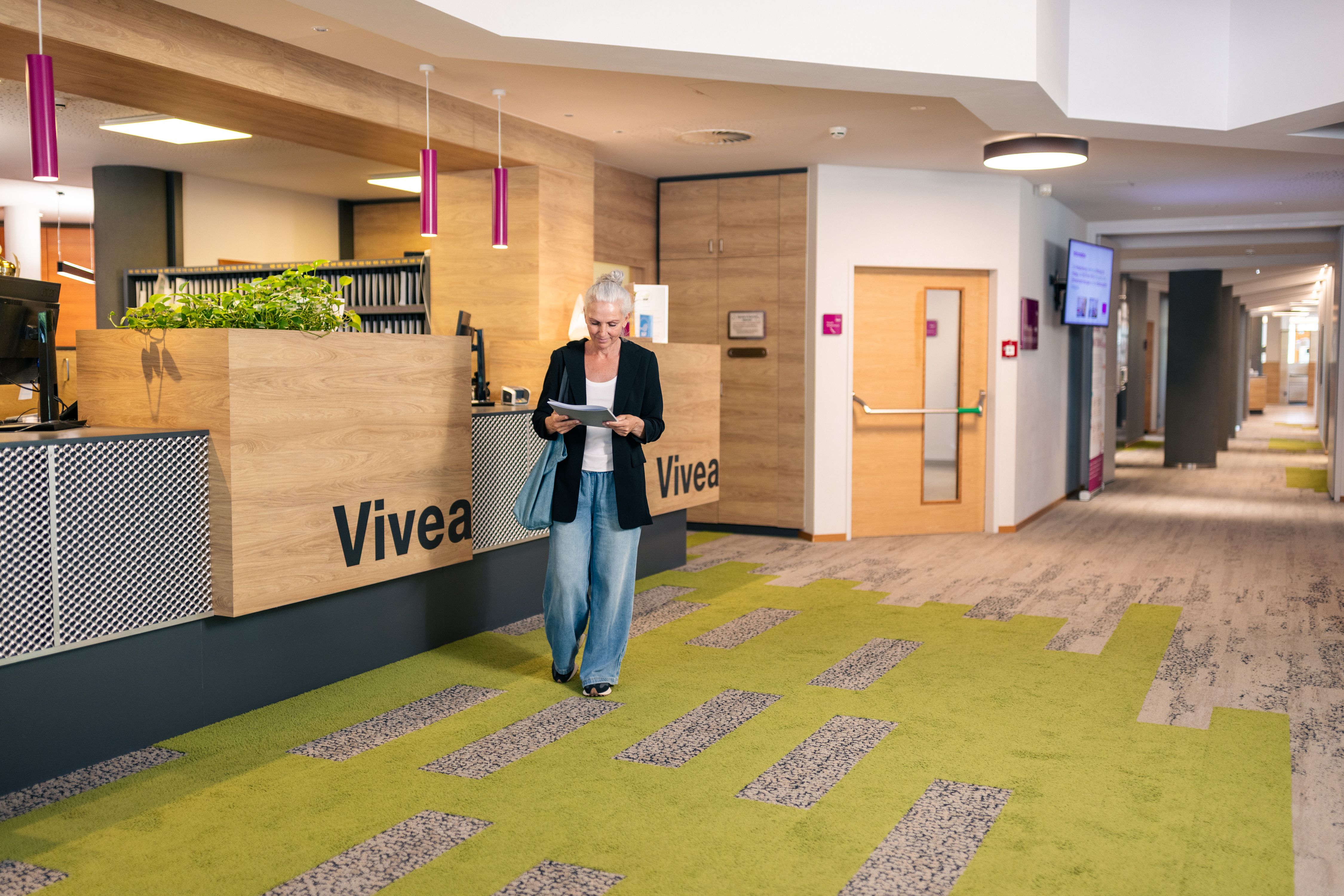 Eine Frau steht in der Lobby des Vivea 4* Hotels Bad Schönau und liest ein Dokument. Die Rezeption ist modern gestaltet mit Holz, Pflanzen und einem grau-grünen Teppich.