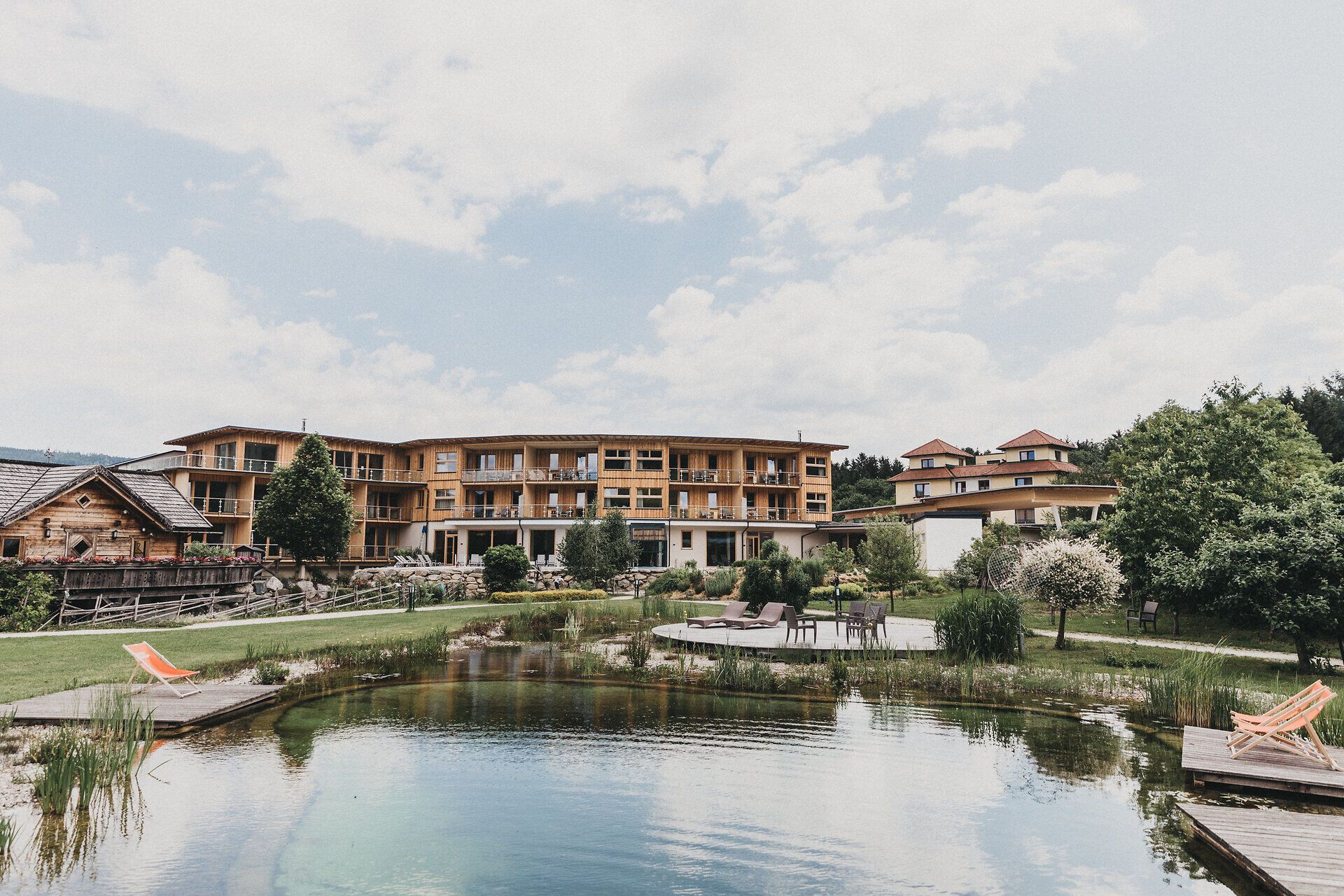 Die Außenansicht eines mehrstöckigen, modernen Hotelgebäudes mit Holzfassade und umlaufenden Balkonen. Im gepflegten Außenbereich befindet sich im Vordergrund ein naturbelassener Badeteich mit Schilfbewuchs. Rechts lädt eine Holzterrasse mit modernen Sonnenliegen und Sitzmöbeln zum Entspannen ein.