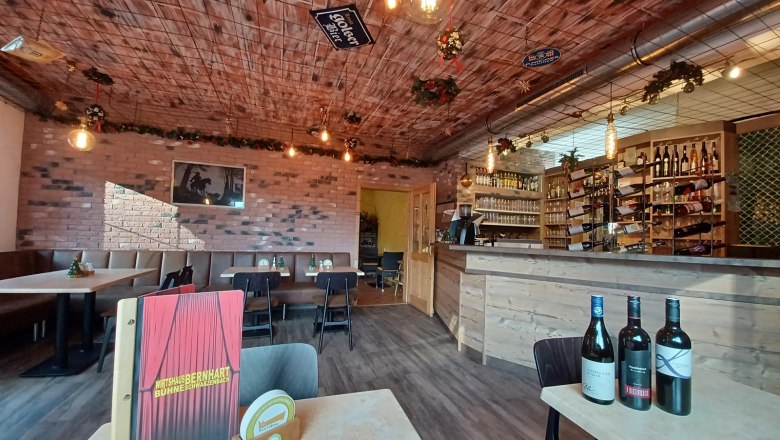 Gemütliche Gaststube mit Holztischen, Bar und Weinflaschen.