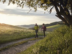Zwei Personen wandern auf einem Feldweg bei Sonnenuntergang in einer h&uuml;geligen Landschaft.