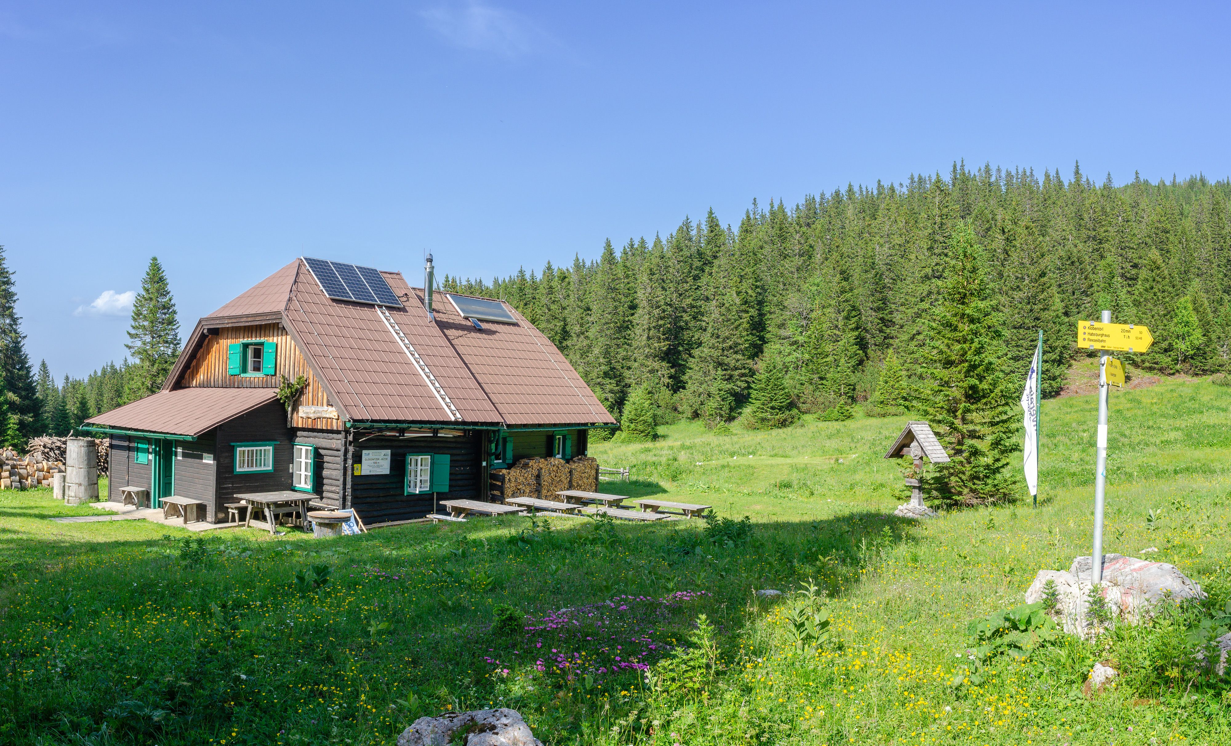 Eine Berghütte mit Solarpanelen auf dem Dach, umgeben von grüner Wiese und Wald.