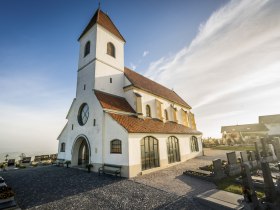 Eine wei&szlig;e Kirche mit rotem Ziegeldach und einem Friedhof im Vordergrund bei sonnigem Wetter.