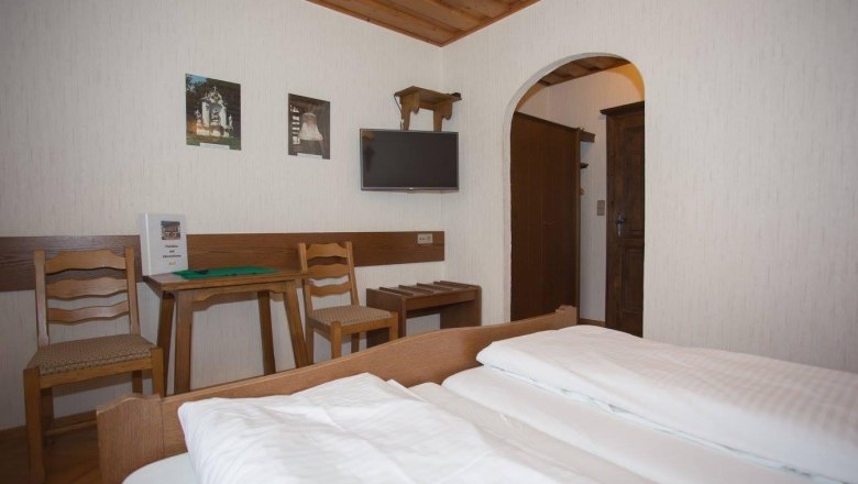 A cozy double room with wooden furniture, a double bed, a small table with two chairs and a TV on the wall.