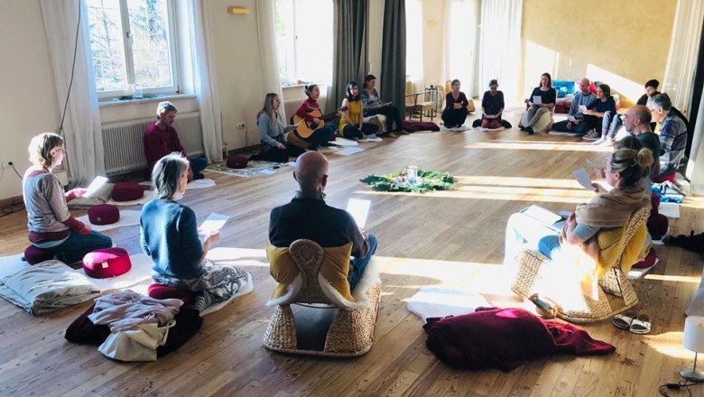 People sit in a circle on cushions and chairs in a bright seminar room with a wooden floor.
