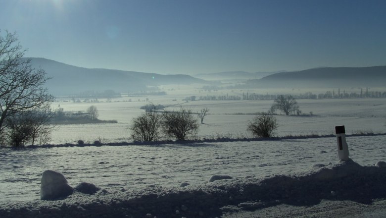 Winterliche Landschaft mit Schnee, Bäumen und Hügeln im Hintergrund, unter strahlender Sonne.
