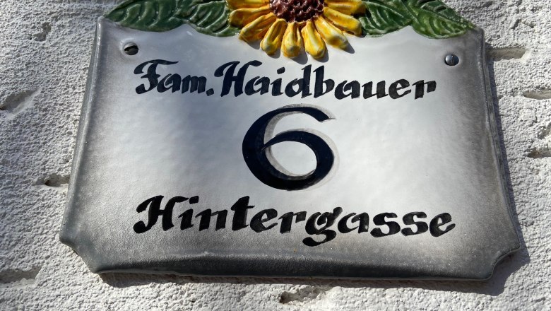 Hausnummernschild mit Sonnenblume und der Aufschrift 'Fam. Haidbauer 6 Hintergasse'.
