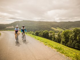 Zwei Radfahrende auf einer kurvigen Stra&szlig;e in einer h&uuml;geligen Landschaft mit der Burgruine Kirchschlag im Hintergrund.