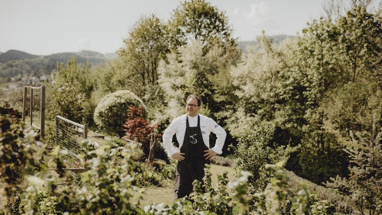 A cook stands in a garden with trees in the background.