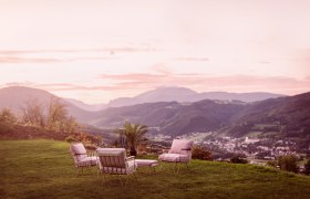 Gartenm&ouml;bel auf einer Wiese mit Blick auf eine Berglandschaft bei Sonnenuntergang.