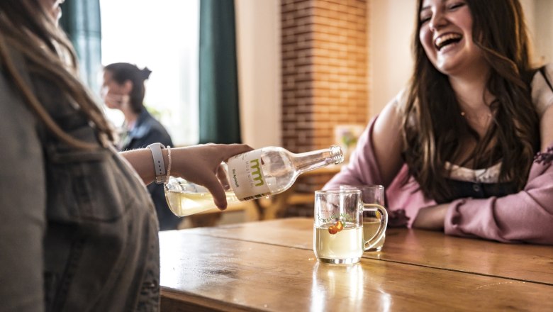 Zwei Frauen sitzen an einem Tisch, eine gießt Most aus einer Flasche in ein Glas. Beide lachen.