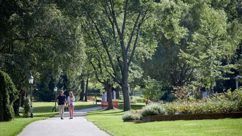 Ein Paar spaziert auf einem Weg im Kurpark Bad Sch&ouml;nau, umgeben von gr&uuml;nen B&auml;umen und gepflegten Blumenbeeten.