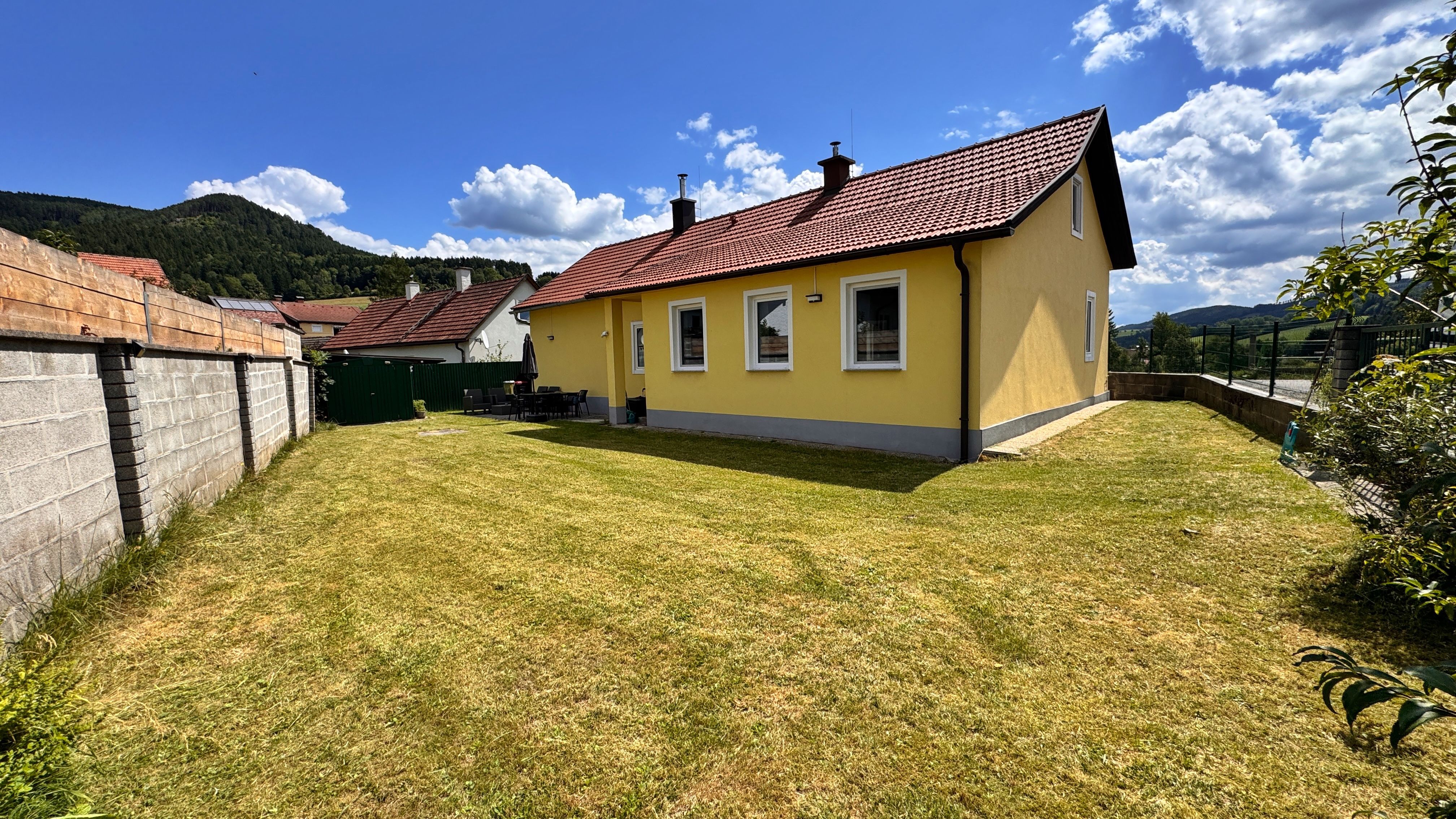Ein gelbes Haus mit rotem Dach in einem Garten mit Rasen, umgeben von einer Mauer und Bergen im Hintergrund.