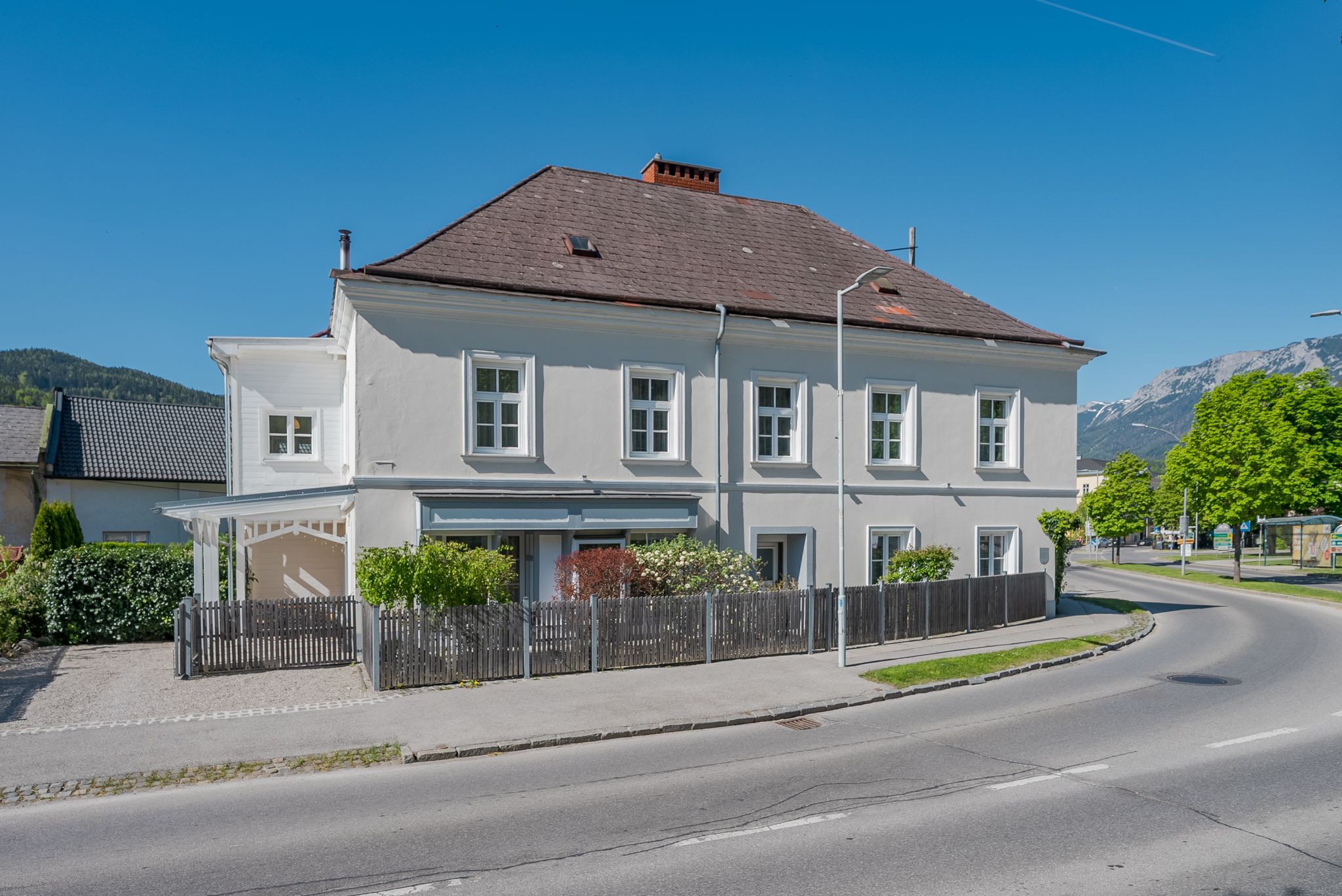 Ein zweistöckiges, weißes Gebäude mit braunem Dach und Holzzaun an einer Straße.