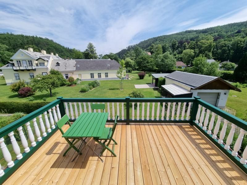 Blick von einem Balkon auf einen Garten mit grüner Wiese und Bäumen, im Hintergrund ein großes Haus und Hügel.