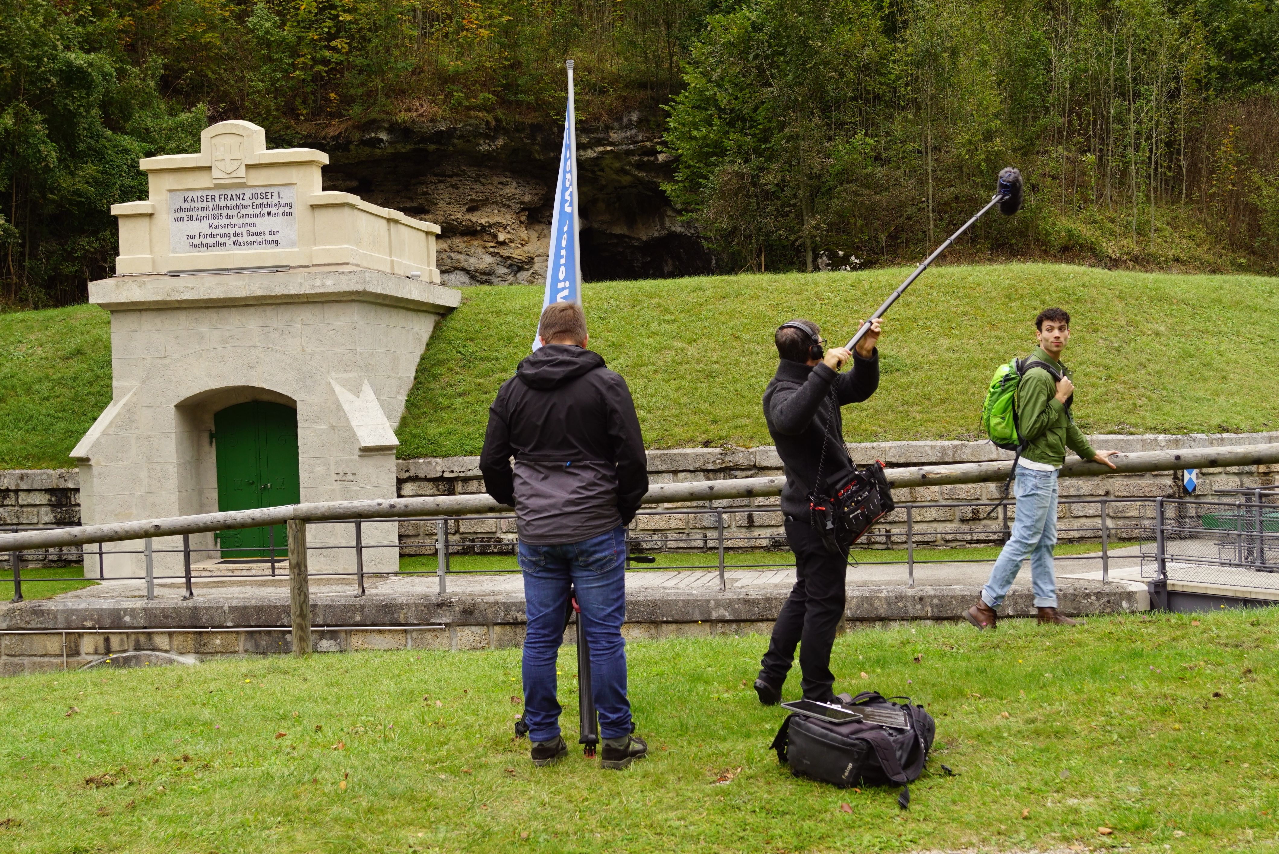 Bei einer Filmaufnahme vor einem steinerner Eingang, in den Hügel gebaut, geht ein Darsteller, der von Kameramann und Tonmann aufgezeichnet wird.