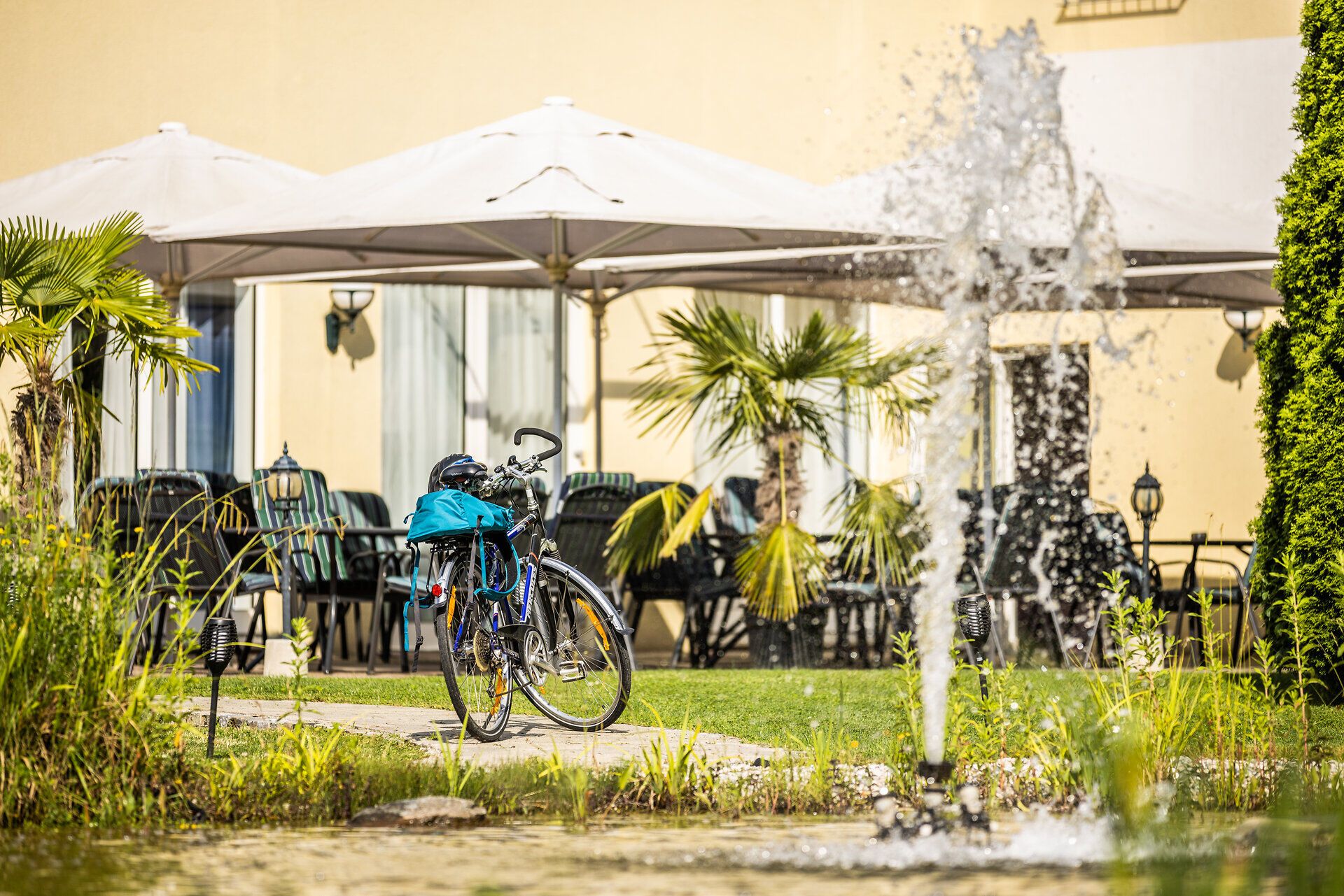 Ein Fahrrad mit Gepäck parkt vor der Terrasse des Grandhotel Niederösterreichischer Hof. Im Vordergrund ist ein Springbrunnen im Garten zu erkennen. 