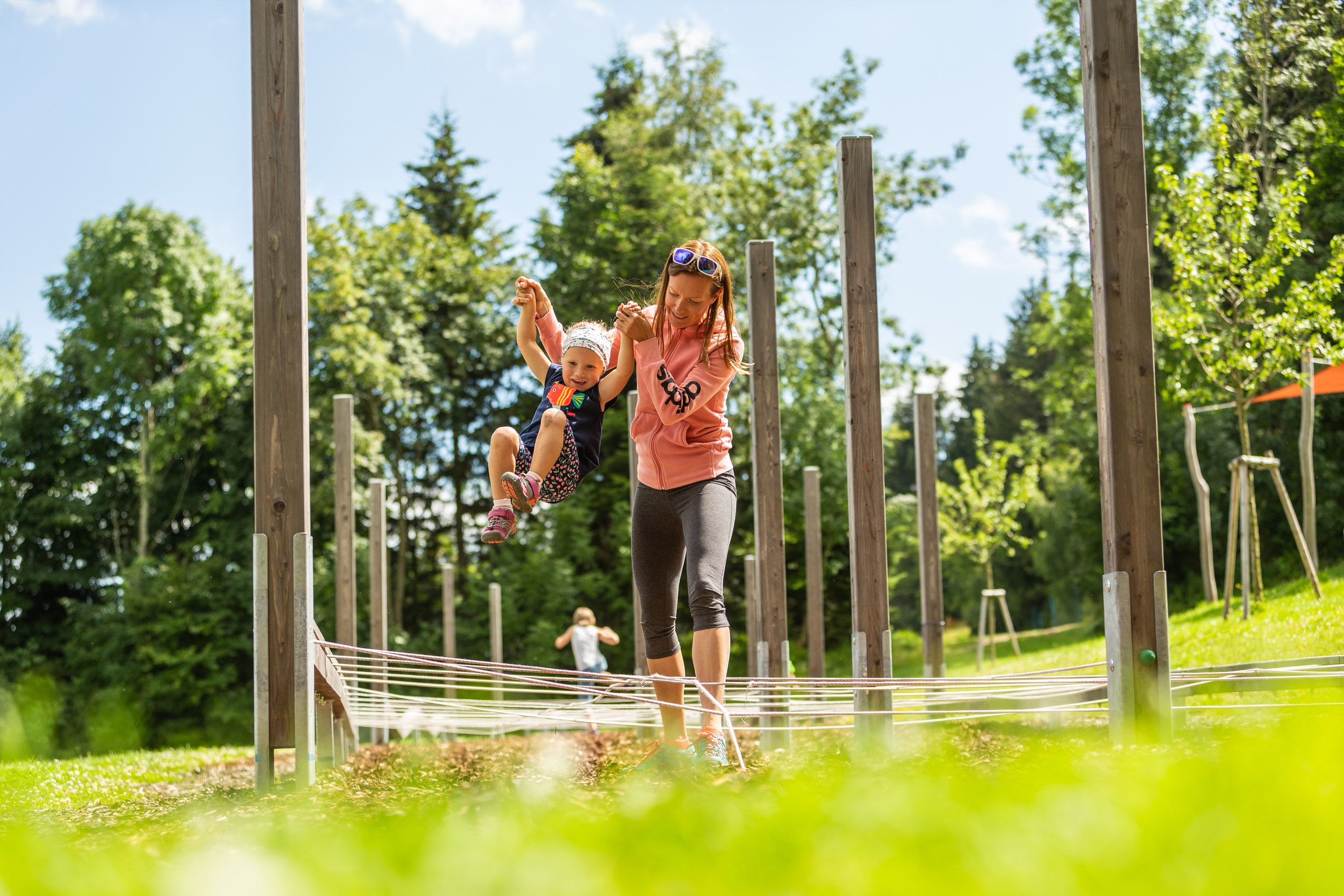 Eine Frau hält ein Kind an den Händen in einem Outdoor-Spielbereich mit Holzpfosten und Seilen.