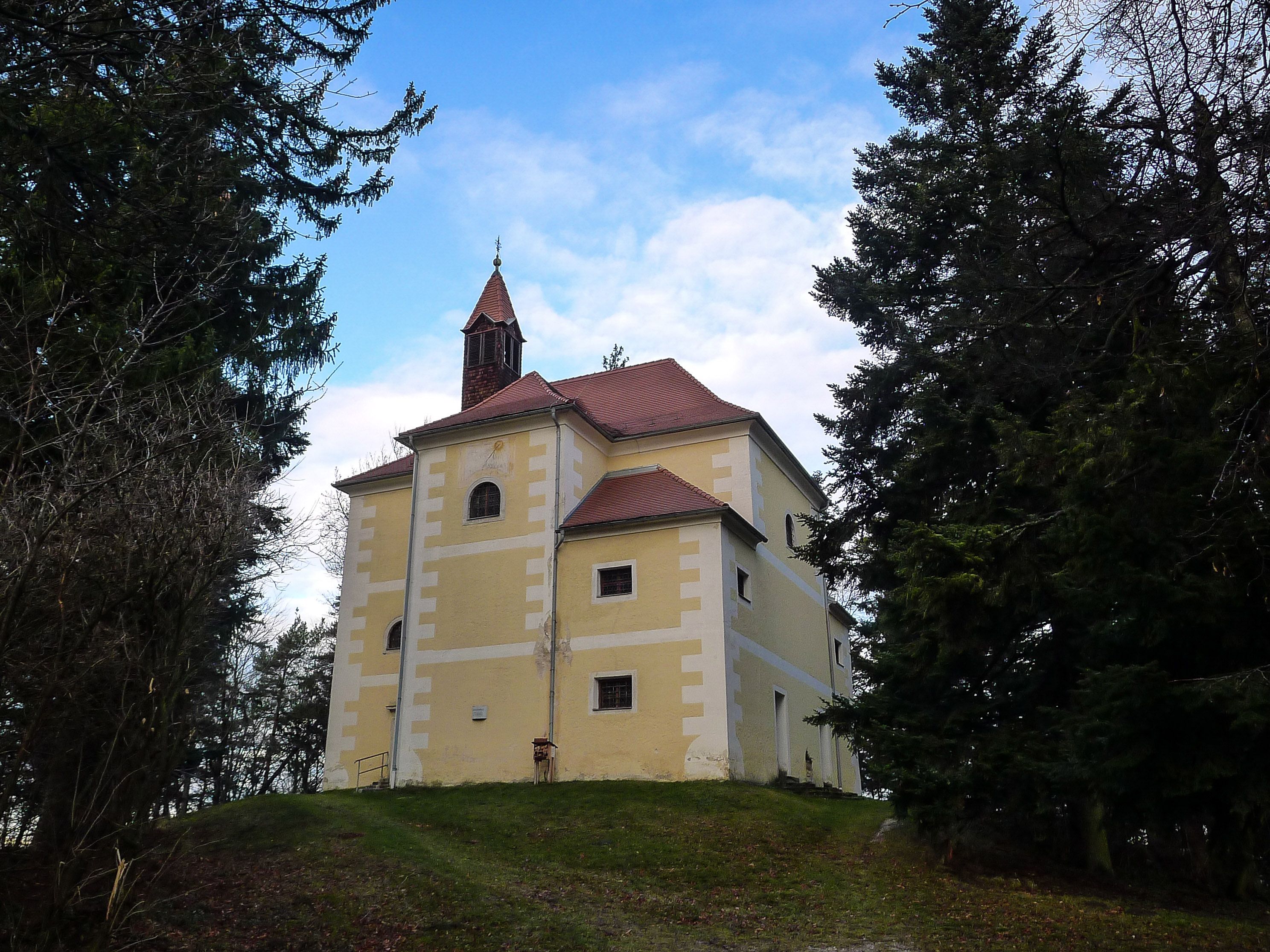 Kapelle mit gelber Fassade auf einem Hügel, umgeben von Bäumen.