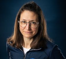 Portraitfoto Wiener Alpen Mitarbeiterin, die eine Jacke mit Wiener Alpen Logo trägt und vor einem dunklem Hintergrund steht.