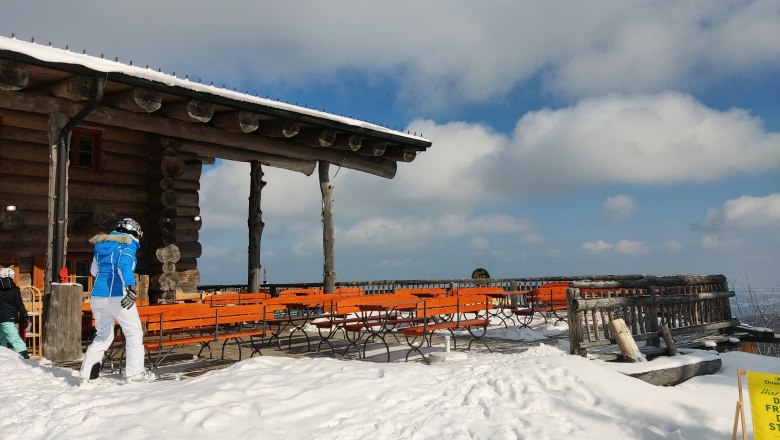 Verschneite Terrasse der Stoa Alm mit Holzbänken und Tischen, blauer Himmel und Wolken im Hintergrund.