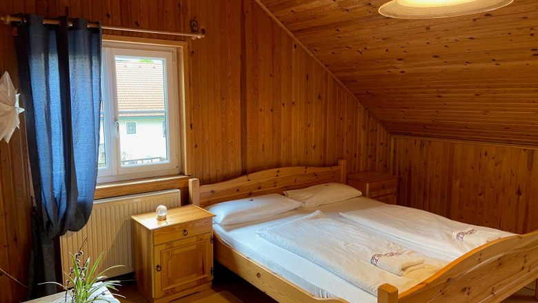 Gemütliches Doppelzimmer mit Holzverkleidung, Doppelbett, Fenster mit blauen Vorhängen und einer Pflanze auf dem Tisch.