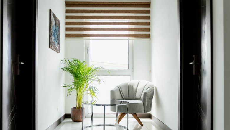 Helle Ecke mit Sessel, Glastisch und Pflanze vor einem Fenster mit Jalousien.