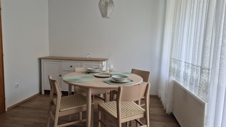 Essbereich mit rundem Tisch, vier St&uuml;hlen und Geschirr, neben einem Fenster mit wei&szlig;en Vorh&auml;ngen.