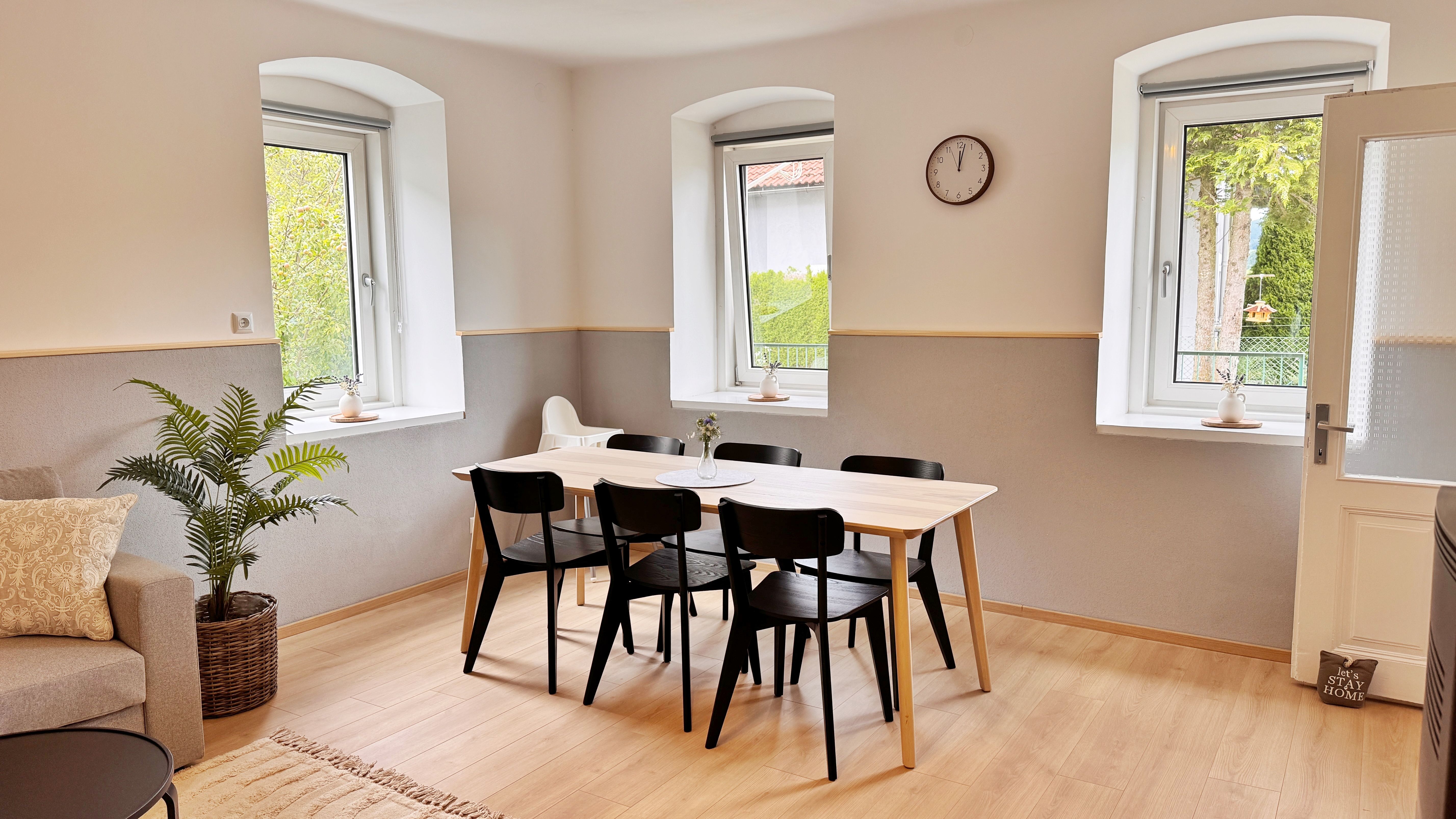 Essbereich mit Holztisch und schwarzen Stühlen, helle Wände, große Fenster, Uhr an der Wand.