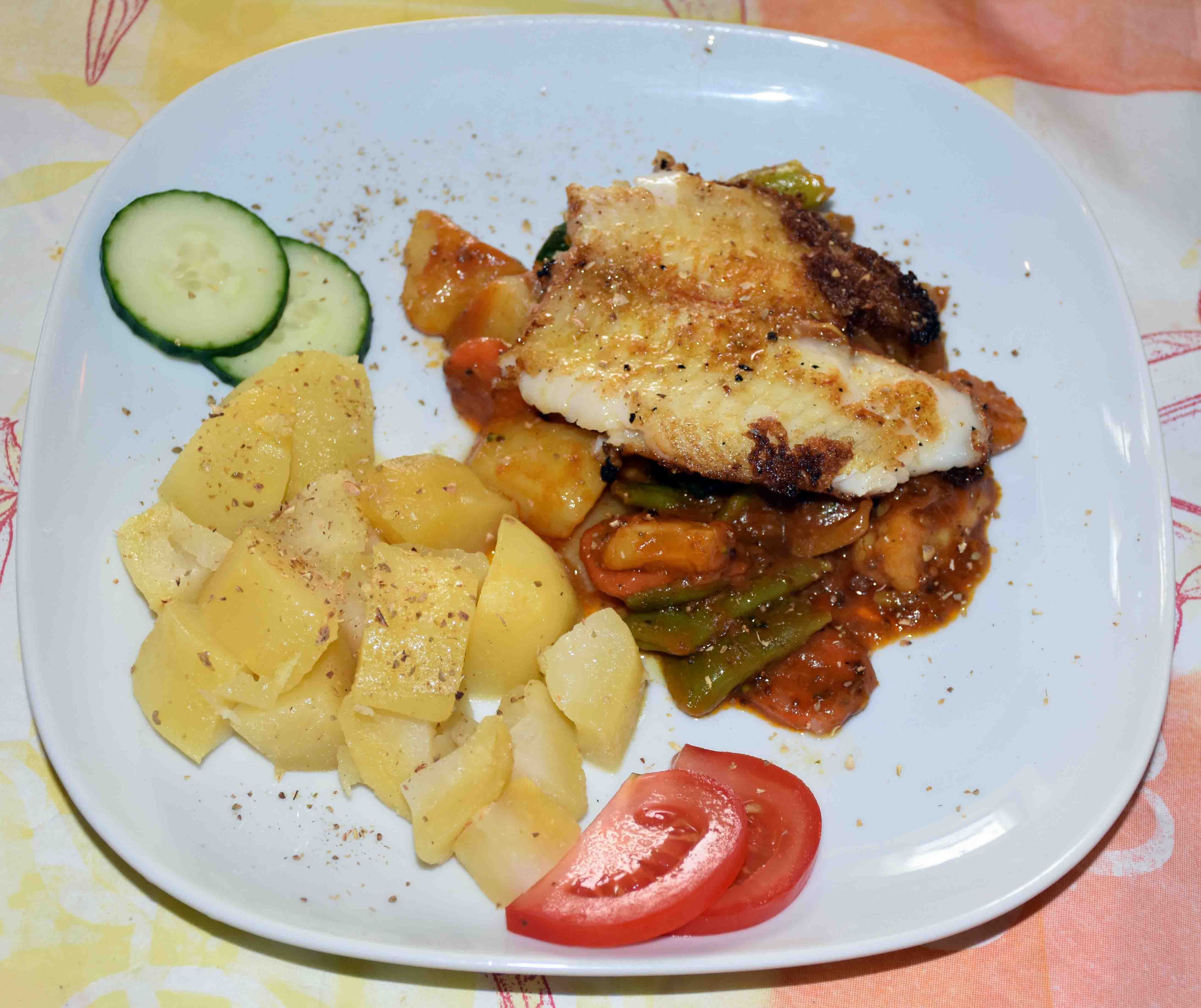 Ein Teller mit gebratenem Fisch, Kartoffeln, Gemüse und Gurken- sowie Tomatenscheiben.