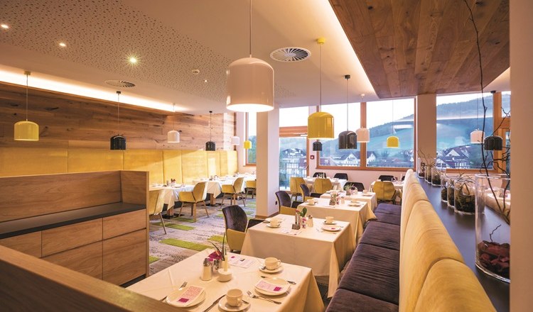 Restaurant mit gedeckten Tischen, Bänken und Stühlen sowie Deckenlampen und großen Fenstern.