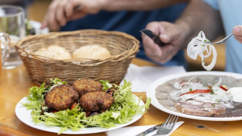 Ein Tisch mit einem Teller voller Fleischlaberl auf Salat, einem Korb mit Gebäck und einem Teller mit Presswurst und Zwiebeln.