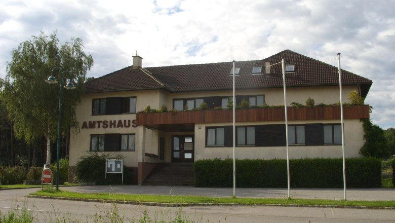 The Natschbach-Loipersbach office building with brown roof and balcony, surrounded by trees and flagpoles.